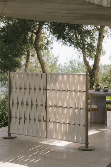 A modern freestanding room divider composed of two rectangular panels. Each panel features a tubular metal frame in a matte taupe color, with horizontal rods through which wide, off-white textile straps are woven in a diagonal, crisscrossing pattern. The divider stands on two flat, circular bases, one at each end. — 3D model