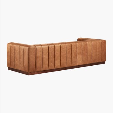 A long, channel-tufted sofa with a minimalist design. The sofa features a continuous padded back and armrest with vertical channels, upholstered in a tan leather-like material. It rests on a low, dark wood plinth base. The overall aesthetic is modern with a touch of art deco. — 3D model