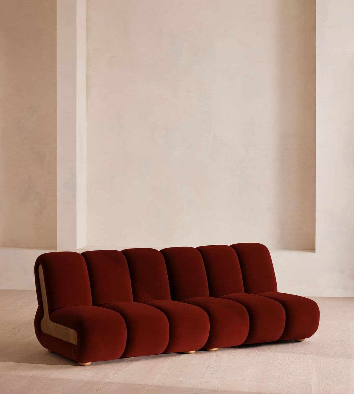 A modular sofa with a distinctive curvilinear design, featuring rounded, segmented cushions for both the seat and back. The sofa is upholstered in a rich, deep red velvet-like fabric. It has a low profile with small, circular wooden feet visible at the base. The armrest on the left side is asymmetrical and integrates a lighter, possibly wood or suede, accent panel. — 3D model