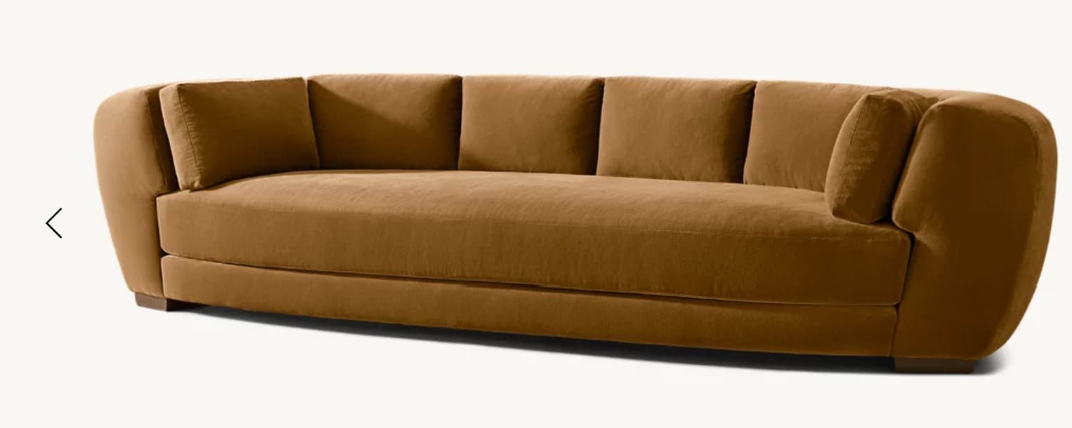 A contemporary, elongated sofa featuring a distinctively curved silhouette. It is upholstered in a rich, warm camel-brown velvet-like fabric, presenting a plush and inviting appearance. The sofa includes deep, individual seat cushions and five separate back cushions, providing ample comfort and support. Its wide, rounded armrests seamlessly integrate with the backrest, contributing to the sofa's organic and fluid form. The design rests on a low-profile base with discreet, recessed wooden feet, creating a perception of floating above the ground. — 3D model