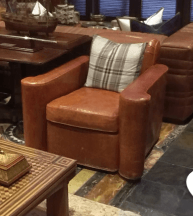 A brown leather armchair with a plaid cushion placed on the seat. The armchair has rolled arms and a slightly distressed leather finish. The backrest is visible, as is part of the seat and the front of the rolled arms. — 3D model