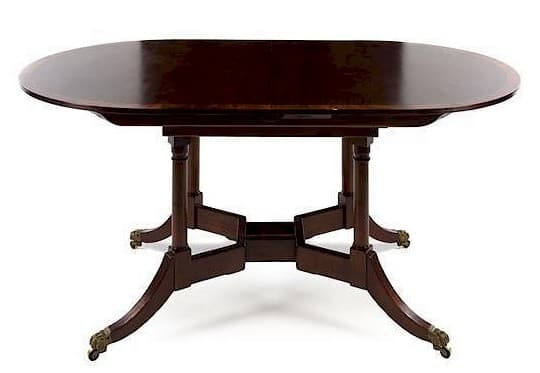 A dark finished, possibly mahogany, dining table featuring an oval top, likely an extension or drop-leaf mechanism, supported by a distinctive double-pedestal base. Each pedestal comprises a central turned support column from which four splayed, saber-style legs radiate. The legs terminate in brass paw-shaped casters, indicating a traditional Regency or Empire period influence. — 3D model