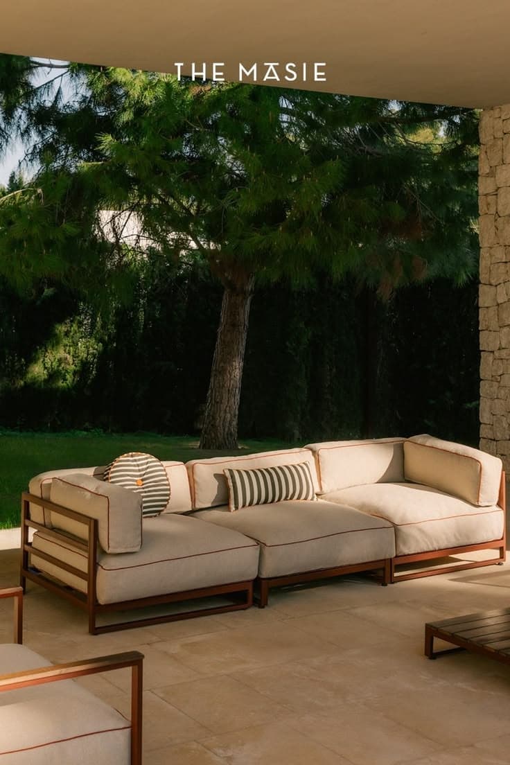 A contemporary modular outdoor sectional sofa featuring a sturdy metal frame and plush, light-colored fabric cushions with contrasting piping. The design includes two main seating modules, an armrest on one end, and comes with two striped accent pillows. — 3D model