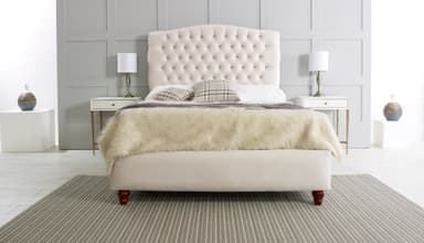 A cream-colored upholstered bed with a diamond-tufted headboard featuring crystal-like embellishments. The bed frame is also upholstered in a matching cream fabric. It is shown with a fluffy cream-colored throw blanket, two plaid throw pillows, and a gray knitted throw. The bed rests on four dark wooden carved legs. Two matching nightstands with drawers and brass accents are positioned on either side of the bed, each holding a white lampshade with a brass base. — 3D model