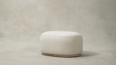A contemporary upholstered ottoman with a rounded oval shape, covered in a plush cream bouclé fabric. The design features soft, curvilinear forms and a low-profile base, contributing to its modern and inviting aesthetic. A subtle seam runs along the perimeter of the top surface, connecting to vertical seams on the sides. — 3D model