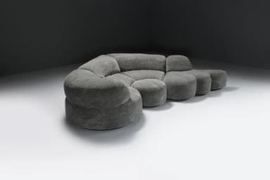 A large, multi-part sectional sofa featuring soft, organic, rounded forms, upholstered in a textured grey fabric. The sofa comprises several modules including corner pieces, armless central sections, and an end module that functions as an extended footrest or chaise. The overall design emphasizes comfort and a flowing, contemporary aesthetic. — 3D model