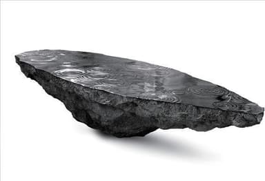 A unique coffee table designed to resemble a floating rock formation with a dark, textured surface, possibly slate or a similar stone, topped with a reflective, water-like surface exhibiting ripple patterns. The underside of the rock shows rough, unworked geological features. The overall aesthetic suggests a naturalistic and artistic design. — 3D model