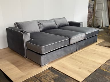 A modern sectional sofa with a chaise lounge extension on one side. The sofa features multiple seat cushions and back cushions, upholstered in a soft, plush gray fabric. It has a piped trim in a lighter contrasting color along the edges of the cushions and frame. Two integrated cup holders are visible on the armrests. The base appears to be a solid structure, possibly with decorative metal feet. — 3D model
