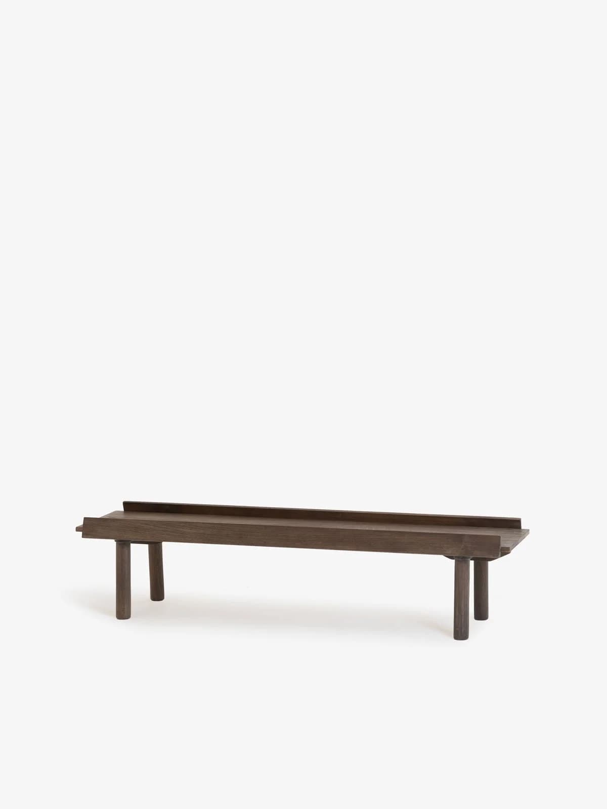 A minimalist wooden bench featuring a long, rectangular top surface with a raised edge along its length, supported by four short, cylindrical legs. The wood has a dark brown finish. — 3D model