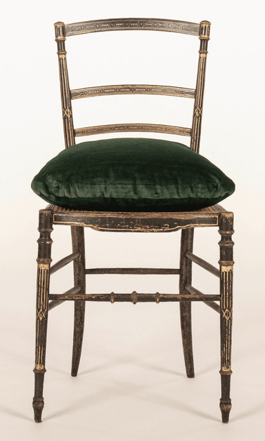 An antique Regency style side chair featuring an ebonized (black lacquered) wood frame with extensive gilt decoration. The chair has a slat back design with three horizontal slats adorned with geometric gilt patterns, complemented by reeded and gilt uprights and finials. The seat appears to be caned, currently topped with a loose, plush dark green velvet cushion. The front legs are turned with gilt accents, while the splayed rear legs are plain. Stretchers connect the legs, some with ring-turned details, and the ebonized finish shows visible distressing and wear. — 3D model