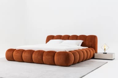 A contemporary platform bed featuring a distinctive design. The headboard and perimeter frame are composed of large, rounded, sausage-like upholstered segments in a warm rust-orange or terracotta fabric. The bed includes white bedding and pillows, presenting a soft, sculptural, and inviting aesthetic. — 3D model