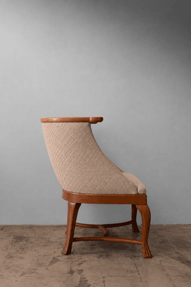 A refined barrel-back armchair featuring a solid wood frame with a polished light brown finish. The chair's back and seat are upholstered in a textured beige fabric with a distinctive diamond quilting pattern. The continuous back curves gracefully to form integrated, low-profile armrests. The chair is supported by four tapered, carved wooden legs, connected by an elegant stretcher base. — 3D model