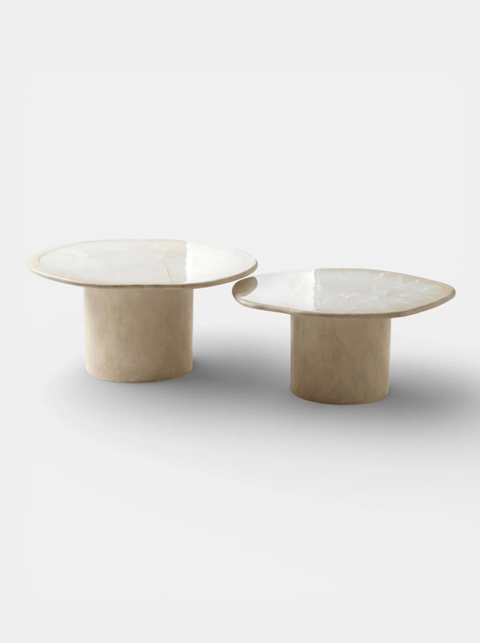 Two low-profile coffee tables are presented. Each table features a wide, flat, irregularly shaped top with a polished, reflective surface exhibiting subtle veining. The tops are supported by thick, cylindrical bases that taper slightly inwards towards the bottom. The overall aesthetic is modern and minimalist. — 3D model