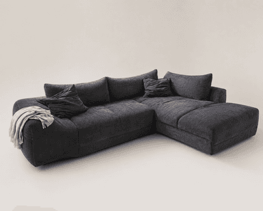 A large, dark gray L-shaped sectional sofa upholstered in a textured fabric, possibly corduroy or chenille. It features multiple plush seat and back cushions, including a prominent chaise lounge section on the right side. Several matching throw pillows and a draped light-colored blanket complete the casual, contemporary look. The sofa has a low-profile design and substantial, inviting seating. — 3D model