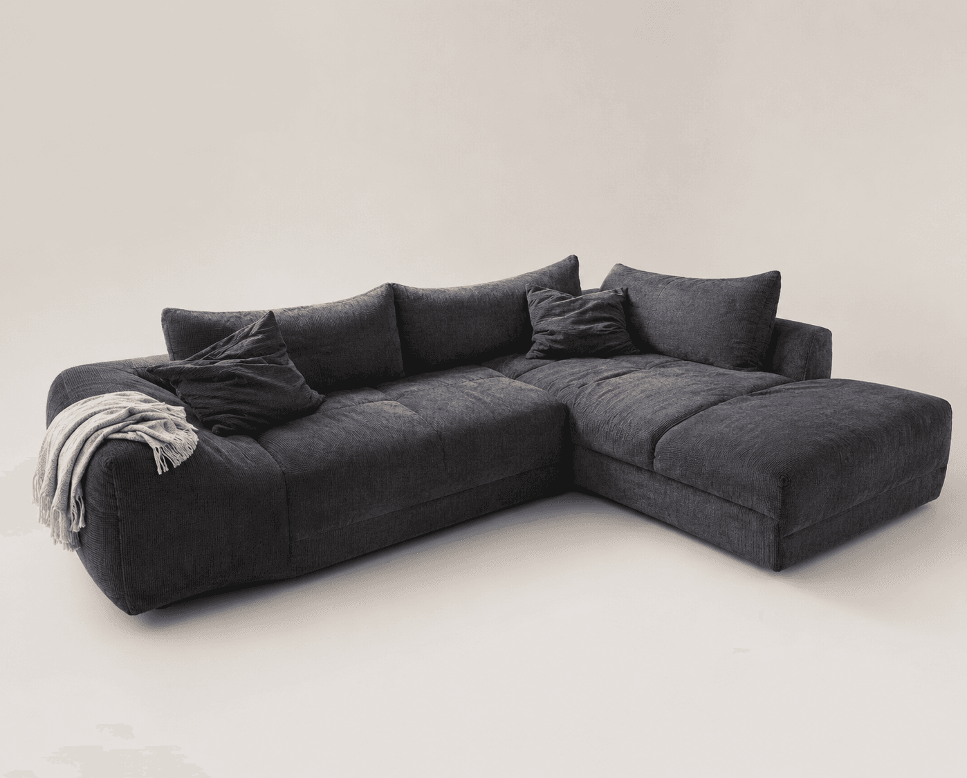 A large, dark gray L-shaped sectional sofa upholstered in a textured fabric, possibly corduroy or chenille. It features multiple plush seat and back cushions, including a prominent chaise lounge section on the right side. Several matching throw pillows and a draped light-colored blanket complete the casual, contemporary look. The sofa has a low-profile design and substantial, inviting seating. — 3D model