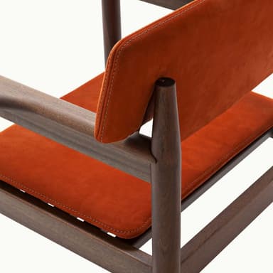 A contemporary armchair featuring a dark wood frame and rust-colored upholstered seat and backrest. The backrest is rectangular with cut-outs to accommodate the rounded wooden armrests, which extend from the rear support posts. The legs are cylindrical and straight, connected by stretcher bars beneath the seat. The seat cushion is subtly tufted. — 3D model