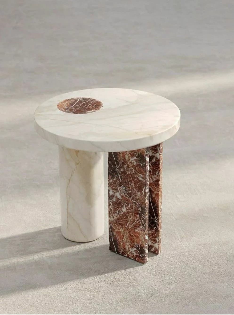 A round side table crafted from two distinct types of marble, featuring a cream-colored top with a recessed circular inlay of reddish-brown marble. The base is composed of an asymmetric arrangement: one cylindrical leg of the cream marble and two flat, rectangular panels of the reddish-brown marble, creating a visually striking and modern design. — 3D model