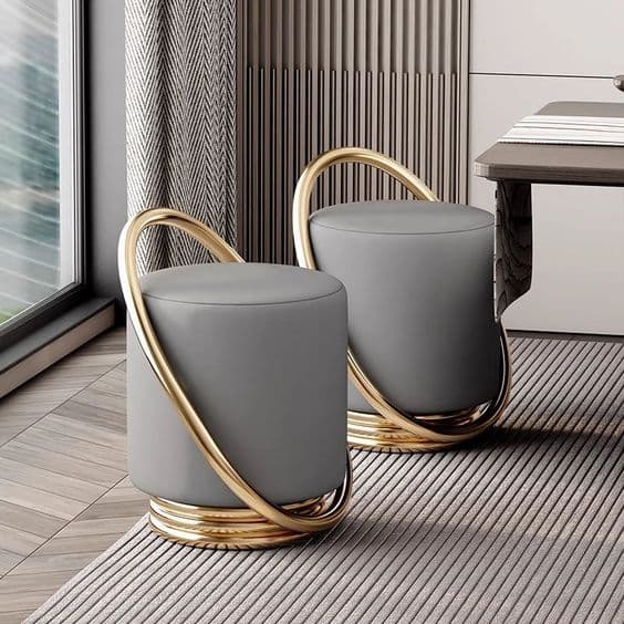 A contemporary cylindrical ottoman featuring soft grey upholstery and a distinctive, curvilinear gold-toned metallic frame that wraps around its body and base. — 3D model