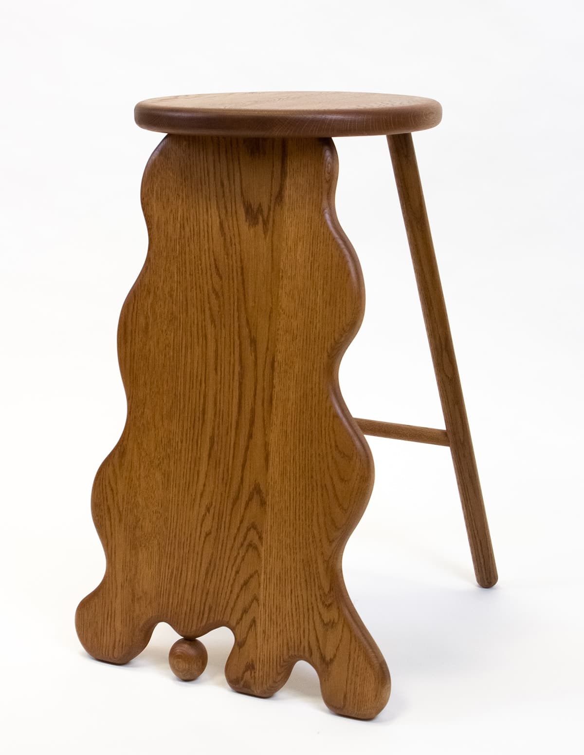 A wooden stool with a distinctive, undulating sculptural base. The seat is round and flat, made of the same wood as the base. The base features a flowing, organic silhouette with rounded extensions, resembling a stylized, abstracted form. One leg is a turned spindle, while the sculptural base provides the primary support. A small wooden sphere is attached to the underside of the sculptural base. — 3D model