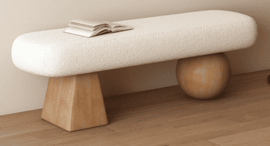 A contemporary bench featuring an elongated, rounded, upholstered seat supported by an asymmetrical wooden base. The seat is covered in a cream-colored bouclé fabric, and the base consists of one triangular, block-like leg and one spherical leg, both made of light-toned wood. — 3D model