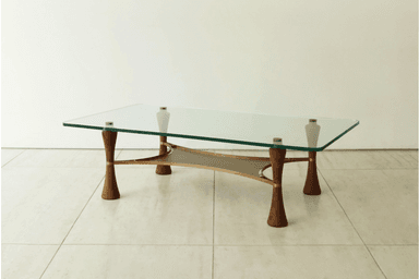A modern coffee table featuring a rectangular clear glass top with softly rounded corners. The base consists of four sculptural, inverted-hourglass-shaped wooden legs, likely walnut, accented with brass rings. These legs support a brass frame that cradles a beige or taupe canvas sling, forming a secondary storage or display surface beneath the glass. The overall design suggests a blend of organic forms and metallic accents. — 3D model