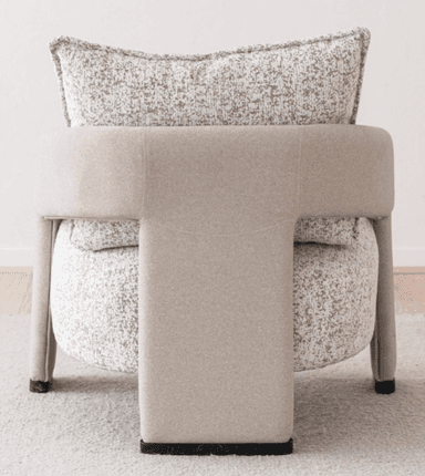 A contemporary upholstered armchair featuring a rounded silhouette and a neutral-toned, speckled fabric. The chair includes integrated, curved armrests and an upholstered backrest, complemented by a separate, plush back cushion with piped edges. The seat appears integrated, and the chair stands on short, upholstered legs or a low, concealed base, suitable for modern interior settings. — 3D model