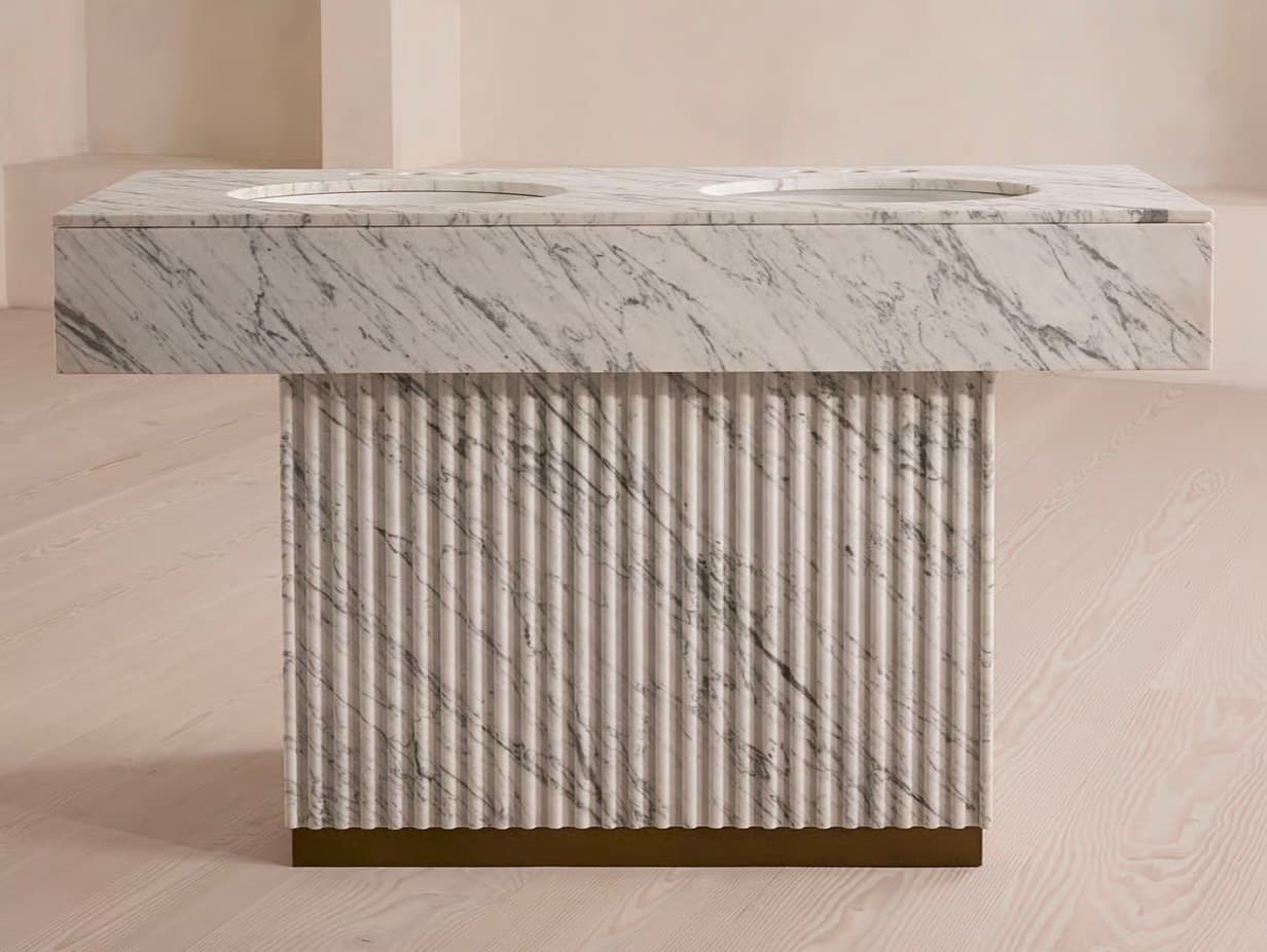 A double vanity with a marble top featuring two integrated sinks. The base is fluted and appears to be constructed from a stone-like material, with a subtle brass accent at the bottom edge. — 3D model