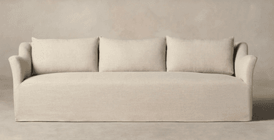 A three-seater sofa, viewed from the rear and side. The sofa features a loose slipcover construction with a skirted base that extends to the floor. It has a low profile with a gently curved armrest visible on the left. The back is composed of multiple cushions, suggesting a comfortable and plush design. The upholstery appears to be a textured linen or linen-blend fabric. — 3D model