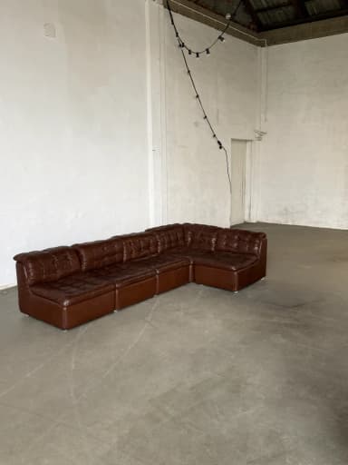 A sectional sofa composed of multiple modular units, upholstered in dark brown tufted material. The sofa forms an L-shape and features a button-tufted design on the seat and back cushions, with clean lines and a low profile. — 3D model