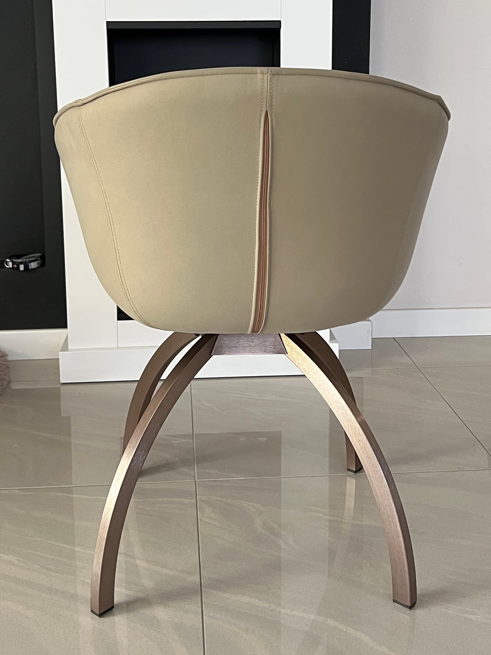 A modern accent chair with a curved, upholstered back and seat. The chair is supported by a distinctive base composed of four curved legs that splay outwards, made of a metallic material. A vertical seam with a zipper-like detail runs up the center of the chair's back. — 3D model