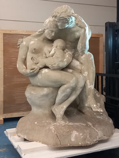 A figurative plaster sculpture depicting a family group. A mother is seated, cradling and breastfeeding an infant, while a father figure leans in from behind, embracing them. The figures are nude, rendered in a naturalistic style, and rest upon an integrated base that shows visible cracks and a distressed, off-white finish. — 3D model