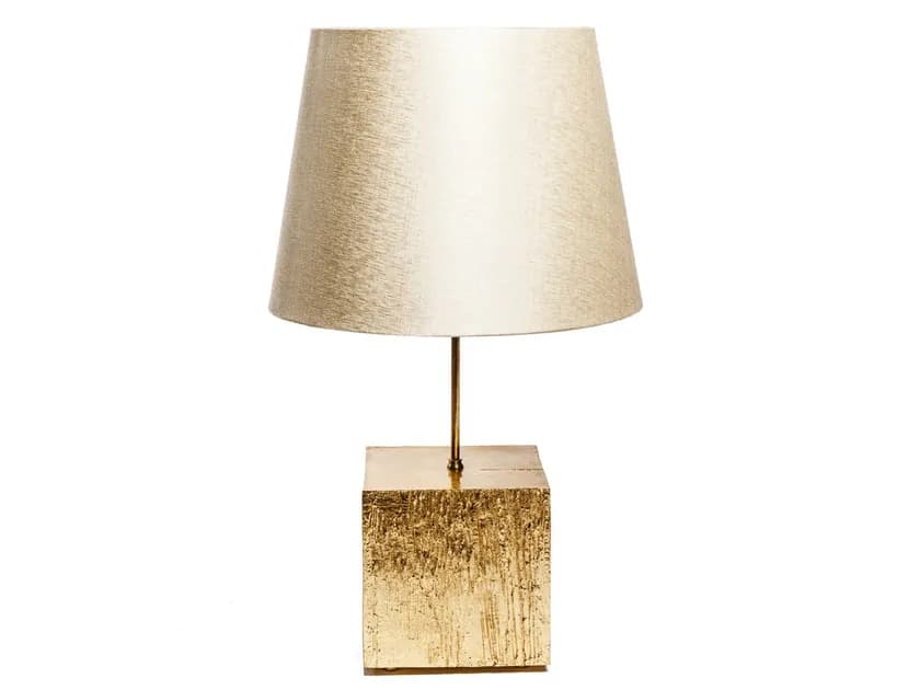 A table lamp with a cube-shaped base and a conical lampshade. The base has a textured, metallic gold finish, appearing to be made of metal with a rough, chiseled surface. A thin, vertical stem connects the base to the lampshade. The lampshade is tapered and has a fabric texture, appearing to be a light, neutral color. — 3D model