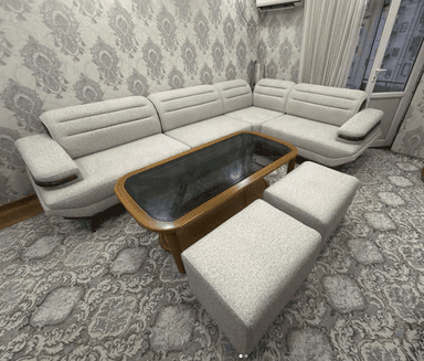 An L-shaped sectional sofa set featuring light gray fabric upholstery, integrated dark wood armrest accents, a rectangular coffee table with a wooden frame and dark glass top, and two matching upholstered ottomans. — 3D model