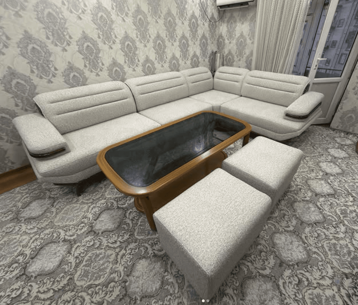 An L-shaped sectional sofa set featuring light gray fabric upholstery, integrated dark wood armrest accents, a rectangular coffee table with a wooden frame and dark glass top, and two matching upholstered ottomans. — 3D model