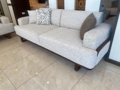 A contemporary two-seater sofa featuring light grey textured fabric upholstery and a visible dark wood frame. The sofa has a low-profile design with integrated backrests and armrests. It includes two back cushions and two decorative throw pillows, one solid brown and one with a black and white geometric pattern. The sturdy wooden base extends along the bottom and curves upwards to support the armrests. — 3D model