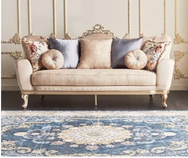 An ornate, gilded chaise lounge upholstered in a beige, velvety fabric. It features a dramatically carved and gilded wooden frame along the backrest and base, with matching cabriole legs. The backrest is asymmetrical, rising higher on one side, and the piece includes a rolled armrest on one end. It is adorned with several decorative throw pillows in varied textures, colors, and patterns, including floral and solid designs. — 3D model