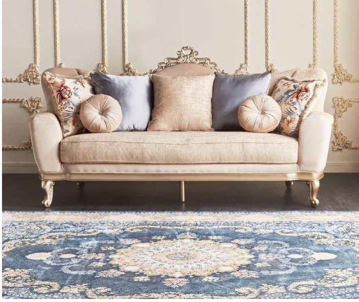 An ornate, gilded chaise lounge upholstered in a beige, velvety fabric. It features a dramatically carved and gilded wooden frame along the backrest and base, with matching cabriole legs. The backrest is asymmetrical, rising higher on one side, and the piece includes a rolled armrest on one end. It is adorned with several decorative throw pillows in varied textures, colors, and patterns, including floral and solid designs. — 3D model