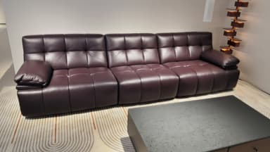 A long, contemporary sofa upholstered in a deep plum-colored leatherette, featuring extensive deep button tufting across its seat, backrest, and low-profile frame. The sofa includes two cylindrical bolster-style armrests, one on each end, which appear to be either adjustable or detachable. The design maintains a minimalist, floor-level profile, emphasizing its plush, segmented appearance. — 3D model