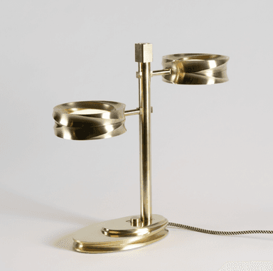A modern two-light table lamp constructed from brass, featuring a central vertical stem with two horizontally extending arms, each supporting a cylindrical light fixture with a distinctive fluted or reeded exterior texture. The base is an oval, layered design with similar fluted detailing. The lamp is illuminated, showcasing integrated LED light sources, and includes a braided fabric power cord. — 3D model