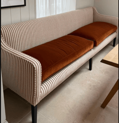 A contemporary banquette-style loveseat with a high, gently curved back and no distinct armrests. It features vertically striped upholstery in beige and brown on the frame, paired with two removable, solid reddish-brown seat cushions. The piece is supported by four slender, tapered dark legs. — 3D model