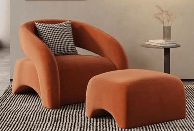A contemporary upholstered armchair featuring a rounded, low-profile design, entirely covered in a rich burnt-orange velvet fabric. The wide armrests and backrest form a continuous, enveloping silhouette, creating a monolithic appearance. A small black and white houndstooth pattern cushion is placed against the backrest. — 3D model