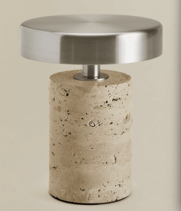 A compact table lamp featuring a cylindrical base crafted from light-colored, porous travertine stone, paired with a flat, circular top made of brushed stainless steel. A thin metallic stem connects the base and the top. The overall design is minimalist and modern, showcasing a contrast in materials and textures. The base has visible natural imperfections typical of travertine, while the metal top has a matte, reflective finish. — 3D model