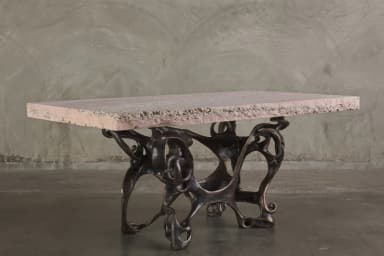 A console table with a rectangular, light pinkish-beige stone top and an intricately sculpted, dark bronze base. The base features an organic, abstract design with interconnected hollow forms and flowing lines, resembling bones or skeletal structures. The stone top has a rough-hewn edge and a textured surface with visible inclusions. — 3D model