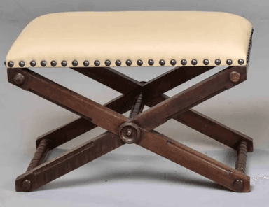 A campaign-style X-frame ottoman featuring a dark wood base and an upholstered seat. The seat is covered in a light, cream-colored material and secured with decorative nailhead trim along the bottom edge. The X-shaped wooden legs are connected by turned stretchers, and the overall design suggests a traditional or vintage aesthetic. — 3D model