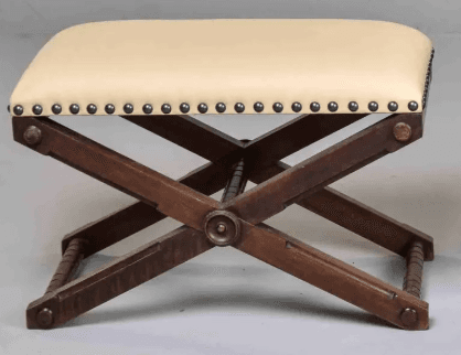 A campaign-style X-frame ottoman featuring a dark wood base and an upholstered seat. The seat is covered in a light, cream-colored material and secured with decorative nailhead trim along the bottom edge. The X-shaped wooden legs are connected by turned stretchers, and the overall design suggests a traditional or vintage aesthetic. — 3D model