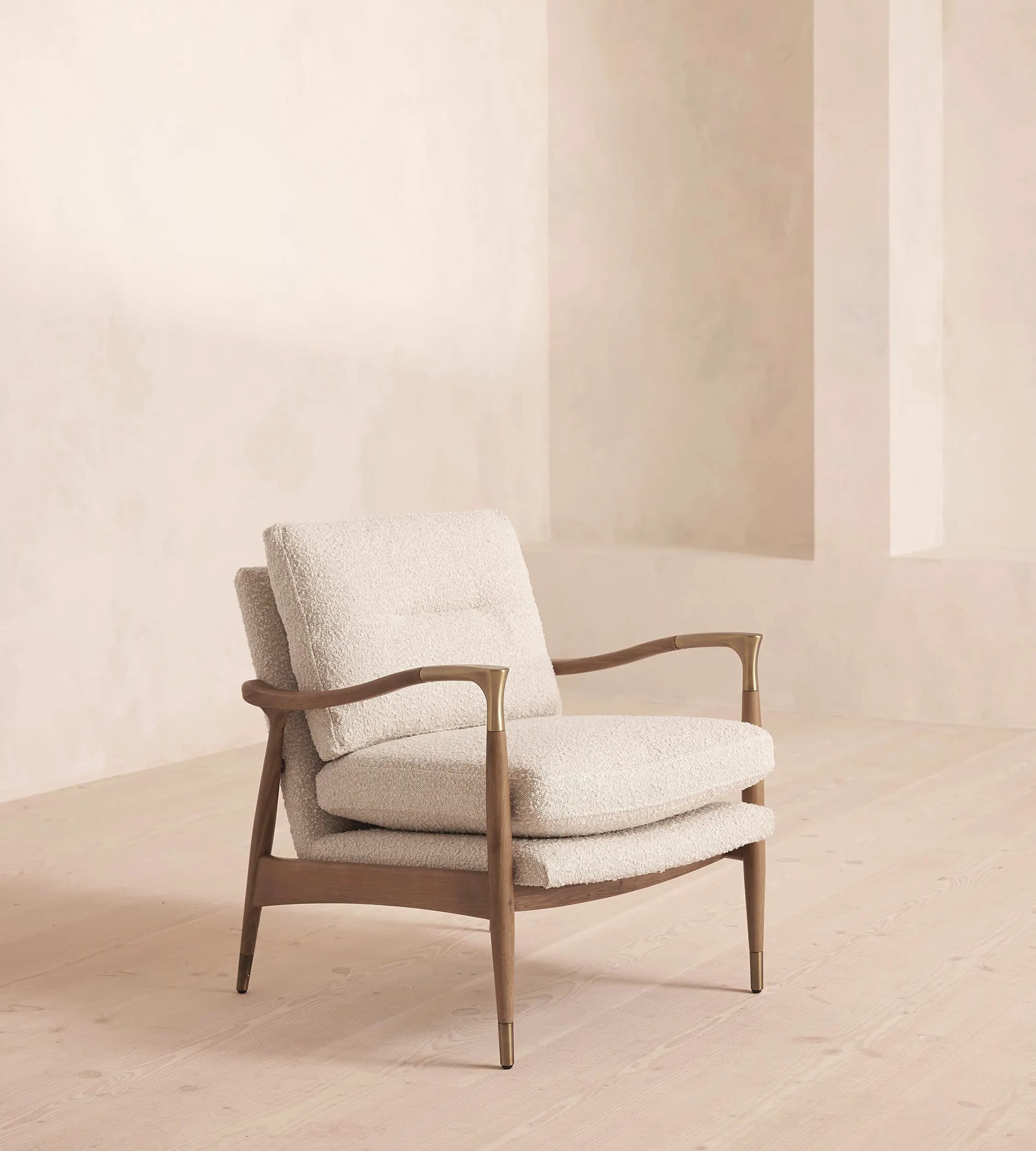 A stylish modern armchair featuring a solid wood frame with a warm finish, complemented by plush bouclé upholstered cushions in a light, neutral tone. The design incorporates elegant curved wooden armrests with subtle brass inlays and tapered legs finished with brass ferrules, giving it a sophisticated and inviting aesthetic. — 3D model