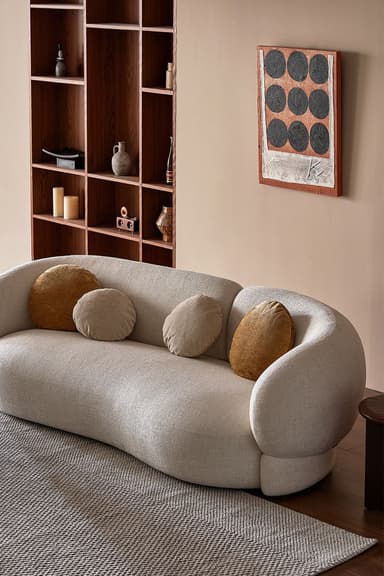 A contemporary curved sofa featuring a fluid, organic silhouette, fully upholstered in a light beige, subtly textured fabric. It has a continuous low-profile back and integrated armrests that seamlessly flow into the seating area. The sofa rests on an upholstered base, slightly elevated from the floor. Four round accent pillows in complementary earthy tones are placed along the backrest. — 3D model