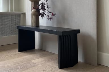 A dark-colored console table with a thick, rectangular top supported by two solid, block-like legs. The legs are set inward from the ends of the tabletop, creating a cantilevered effect. The front edges of the legs have a subtle vertical fluting or grooved detail. The overall design is minimalist and modern. — 3D model