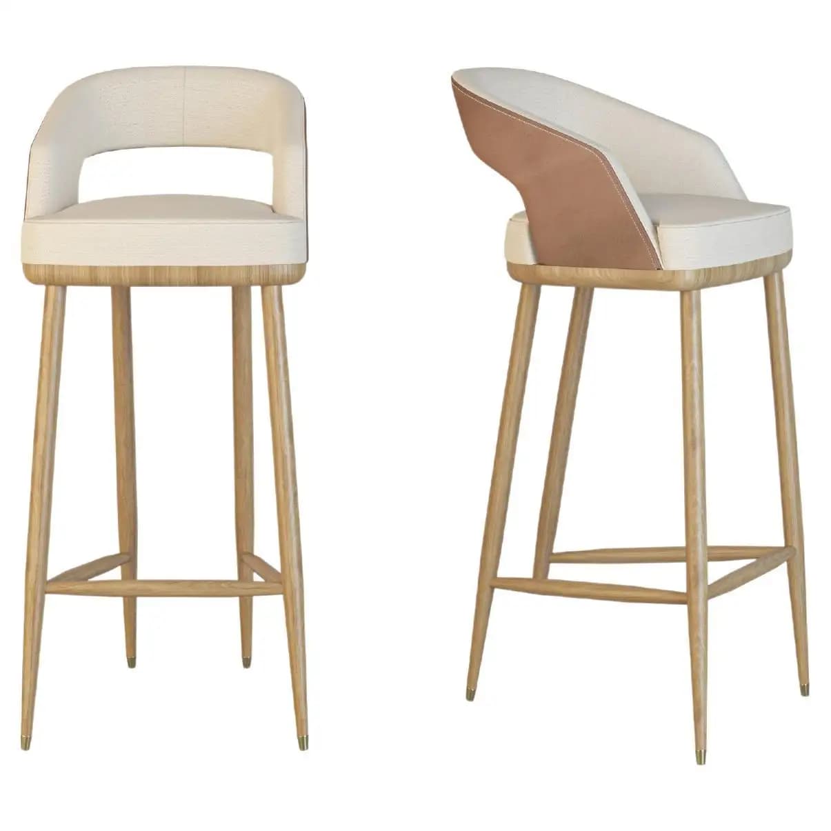 A pair of modern upholstered bar stools featuring a two-tone design with a cream fabric seat and back, complemented by a tan faux leather accent on the exterior of the backrest. The seat and backrest are rounded with integrated, curved armrests and an open cut-out in the lower back. The stools are supported by four tapered light-toned wooden legs, connected by a rectangular footrest, and finished with small brass caps on the feet. — 3D model