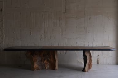 A large dining table with an irregular, organic-shaped top and two substantial, textured bases that resemble natural rock formations or tree trunks. The tabletop appears to be made of multiple panels, possibly wood or stone, with a dark, possibly metal or resin, edge banding. The bases are dark brown and have a rough, sculpted texture, giving the impression of raw, natural elements. — 3D model